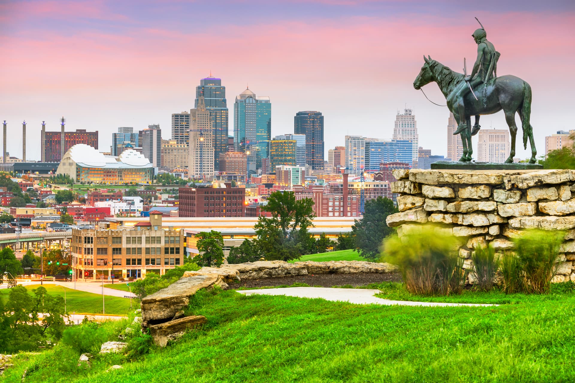 Kansas City skyline at sunset with The Scout statue overlooking downtown from Penn Valley Park, pink sky and green foreground