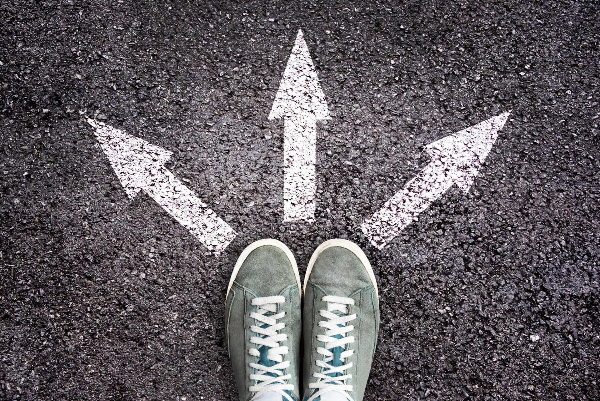 Green sneakers standing at three white directional arrows painted on asphalt, pointing left, forward, and right