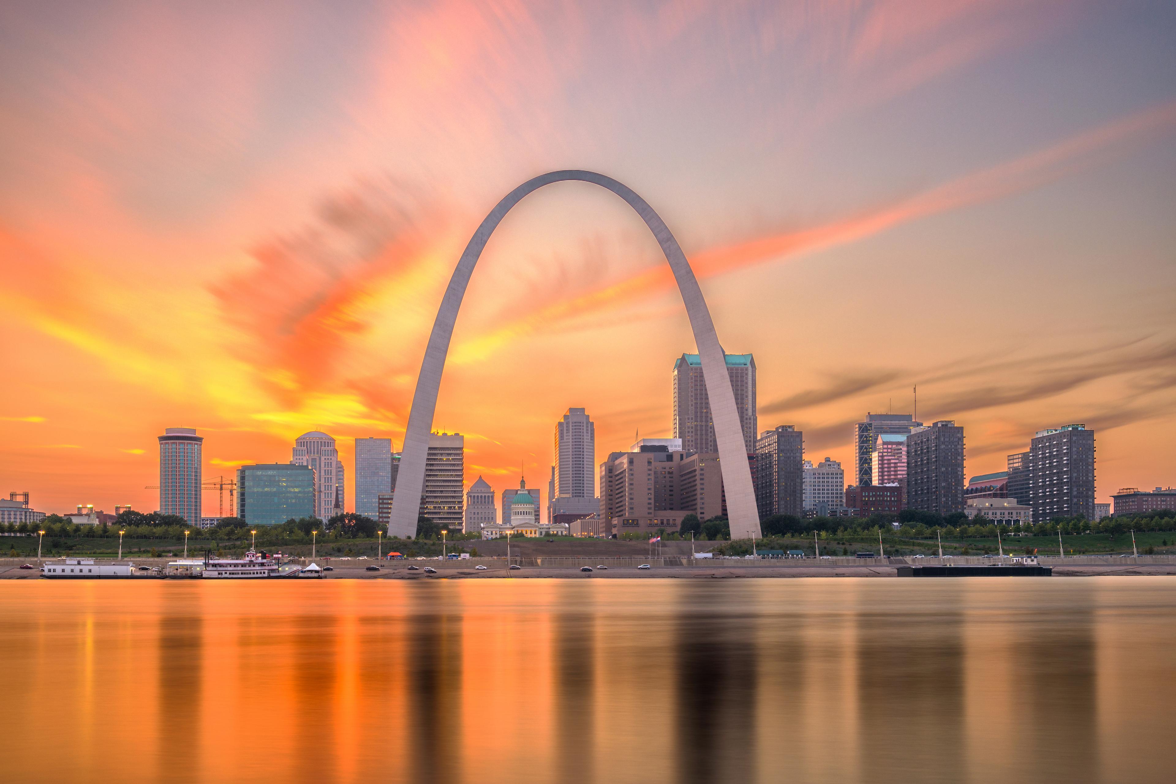 Gateway Arch and St. Louis skyline silhouetted against a dramatic orange sunset sky, reflected in the Mississippi River