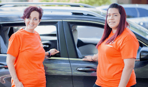 Two team members wearing bright orange polo shirts standing by a black car in a parking lot