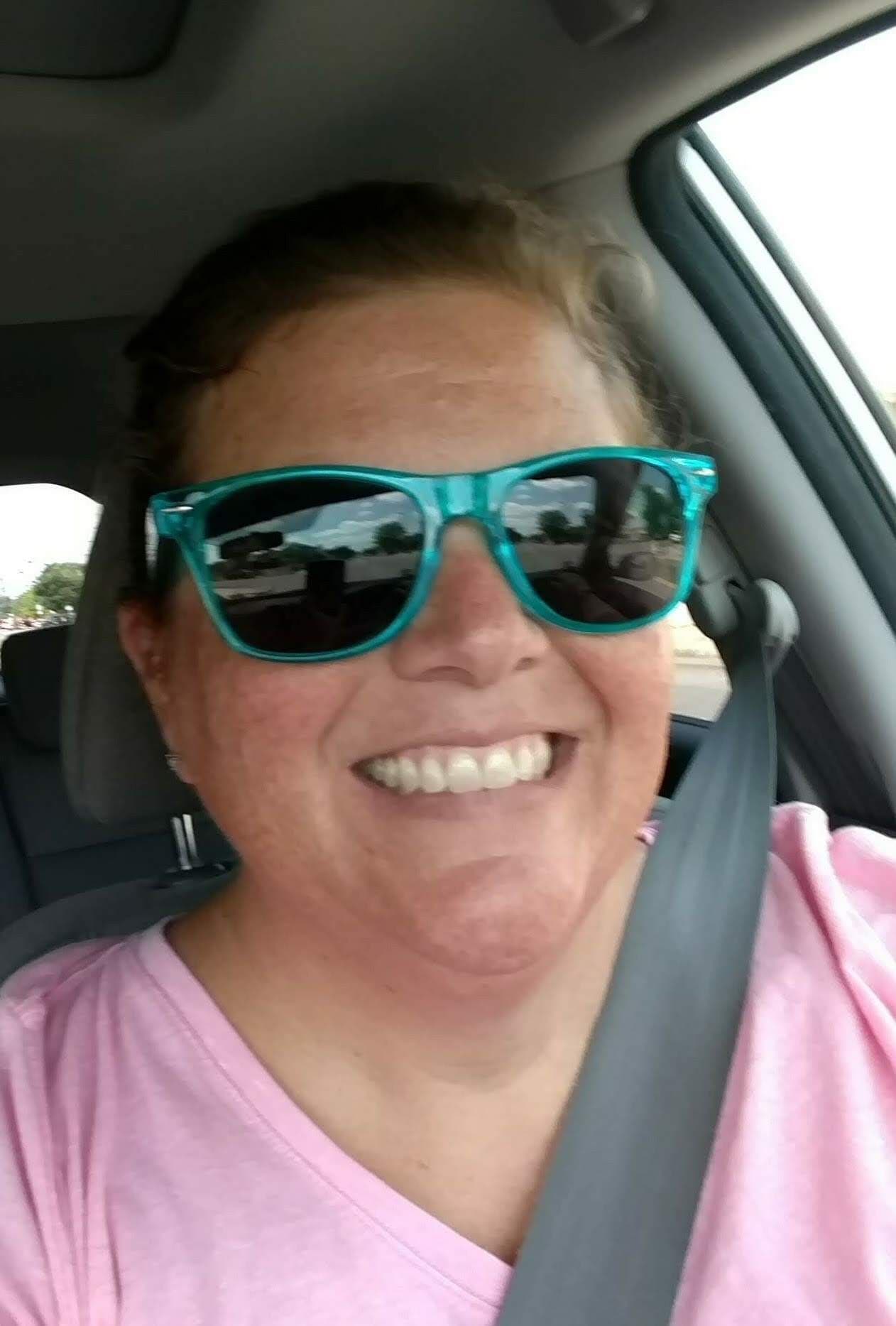 Person wearing bright turquoise sunglasses and pink shirt smiling broadly in car selfie