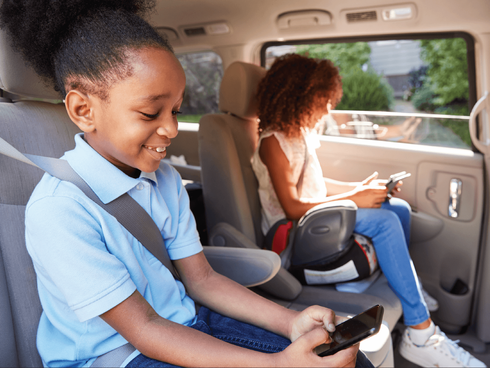 Two children sitting in car backseat with seatbelts on, smiling while using mobile phones