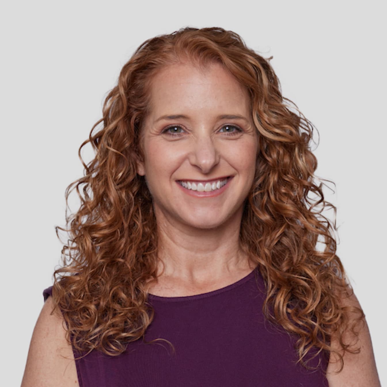 Professional headshot of a person with long curly auburn hair wearing a purple sleeveless top, smiling warmly against a light background