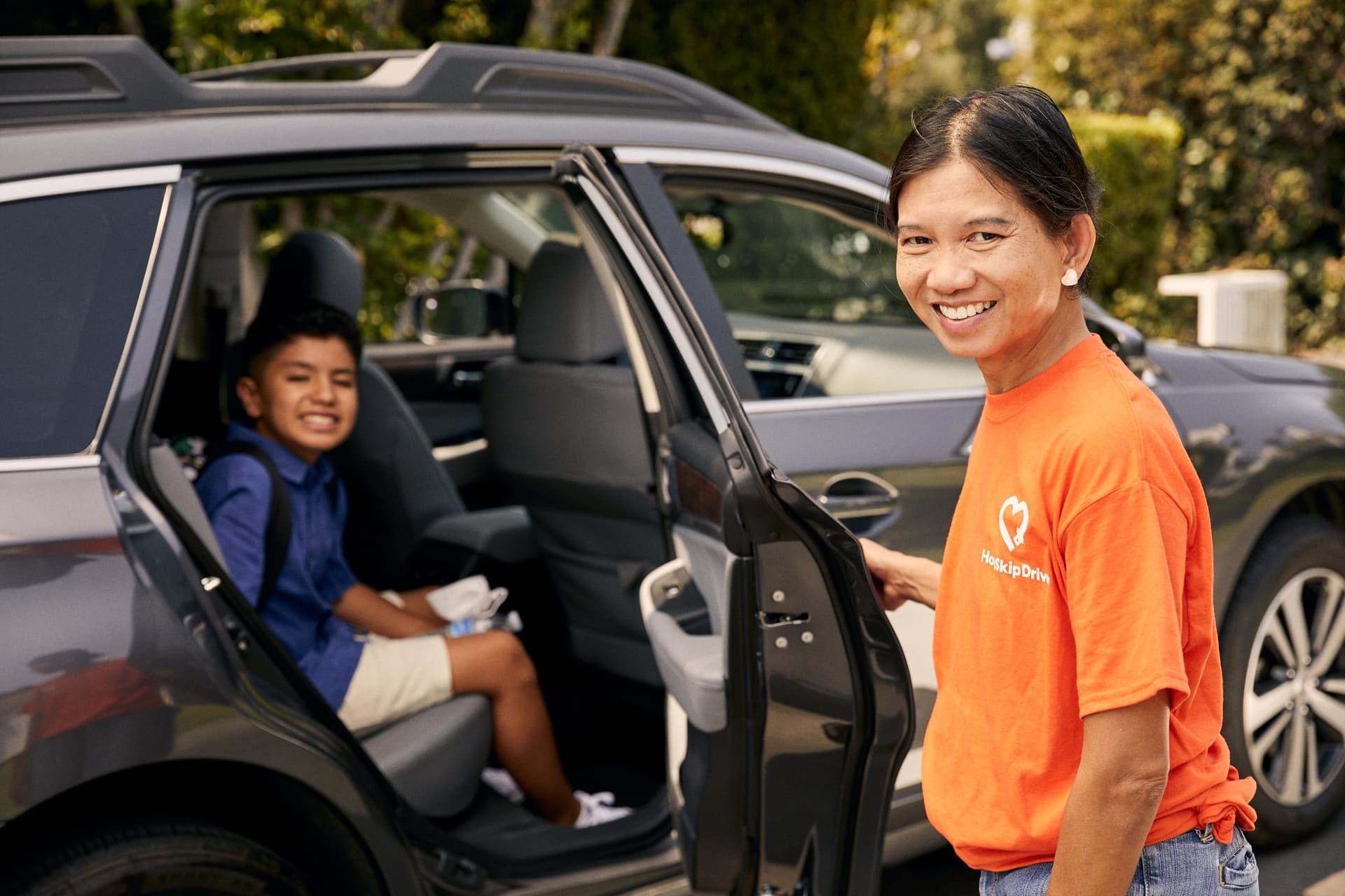 Driver in orange HopSkipDrive shirt opens car door for young student passenger in school uniform