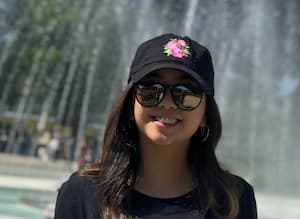 Woman wearing black baseball cap and sunglasses smiling in front of a water fountain on a sunny day