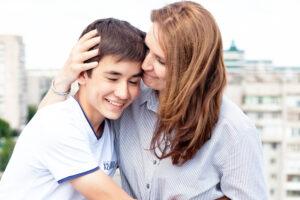 Mother and teenage son sharing an affectionate moment outdoors, both smiling warmly with urban backdrop