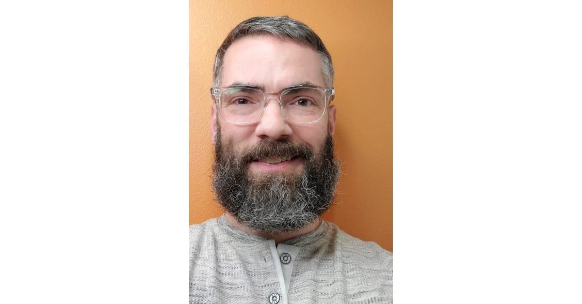 Professional portrait of a smiling man with a full grey beard wearing clear-framed glasses against an orange wall