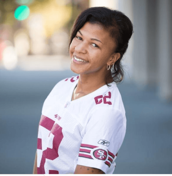 Woman wearing white San Francisco 49ers jersey smiling warmly at camera in outdoor setting with blurred background