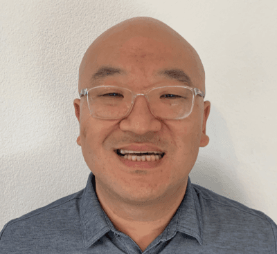Professional headshot of a person wearing clear-frame glasses and a grey collared shirt, smiling warmly against a white background
