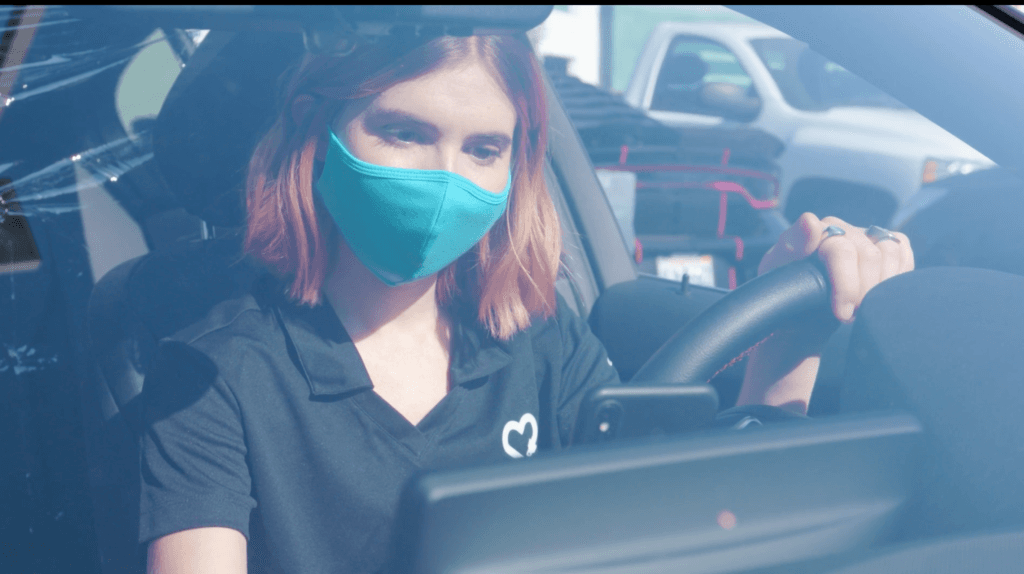 Driver wearing teal face mask and black uniform shirt with heart logo, sitting behind wheel of car