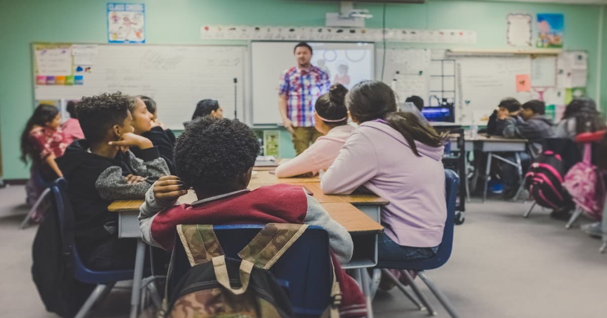 Students sitting at desks listening to teacher in front of whiteboard in modern classroom with mint green walls and educational posters
