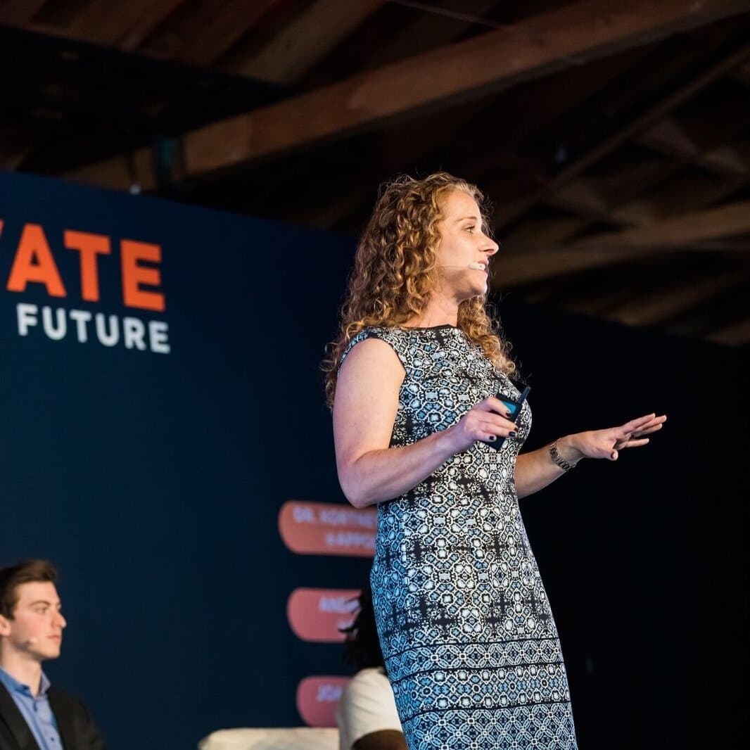 Speaker in patterned dress giving presentation on stage with 'Future' text visible on background screen