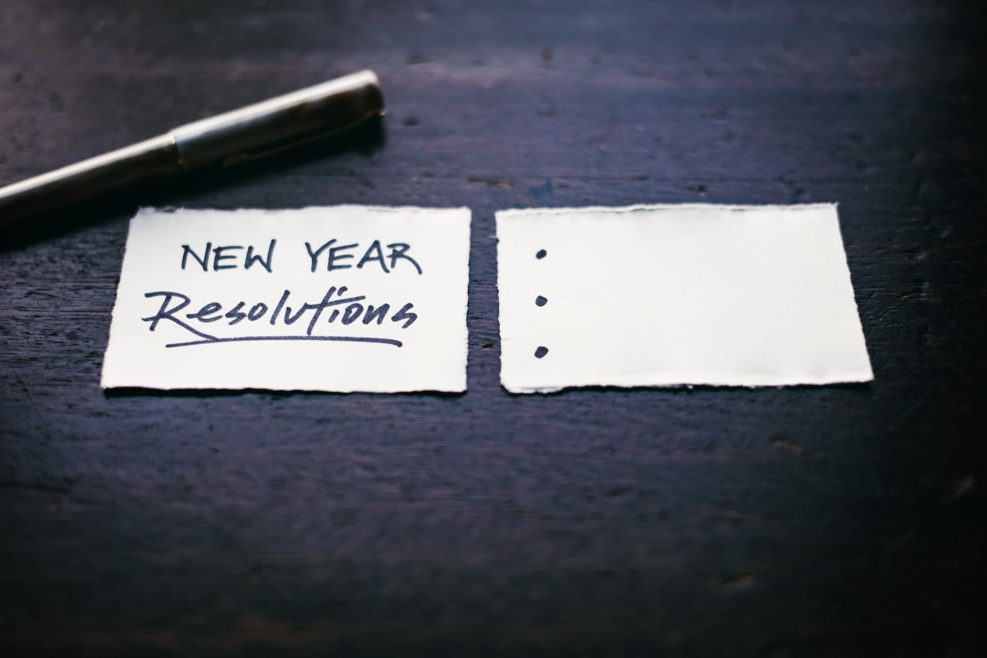Two torn paper pieces on dark surface - one reads 'New Year Resolutions' with bullet points on blank second piece
