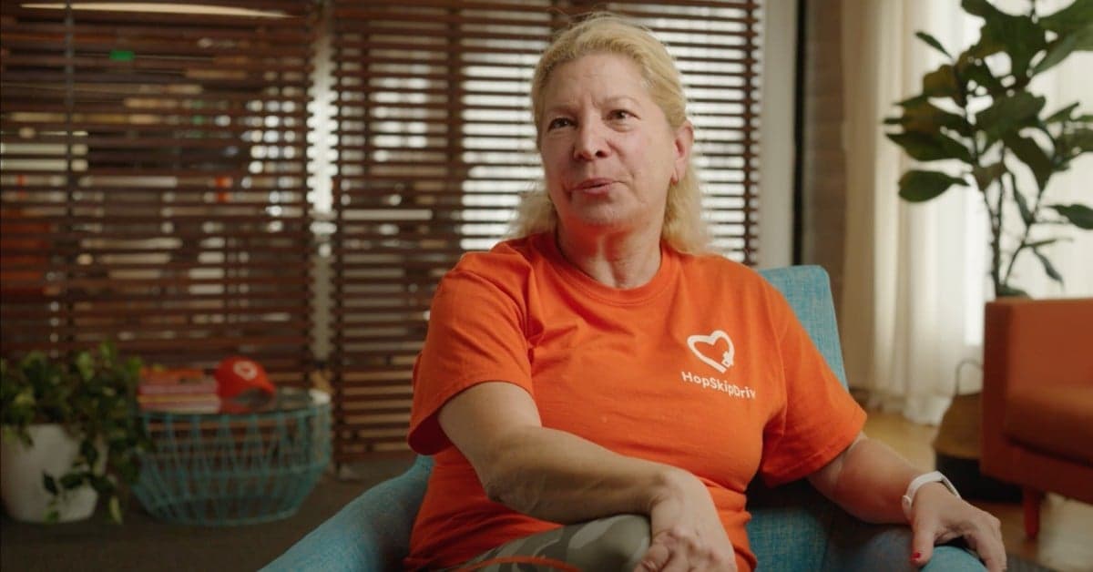 Woman in bright orange HopSkipDrive t-shirt sitting in blue chair during interview, home or office setting with plants in background