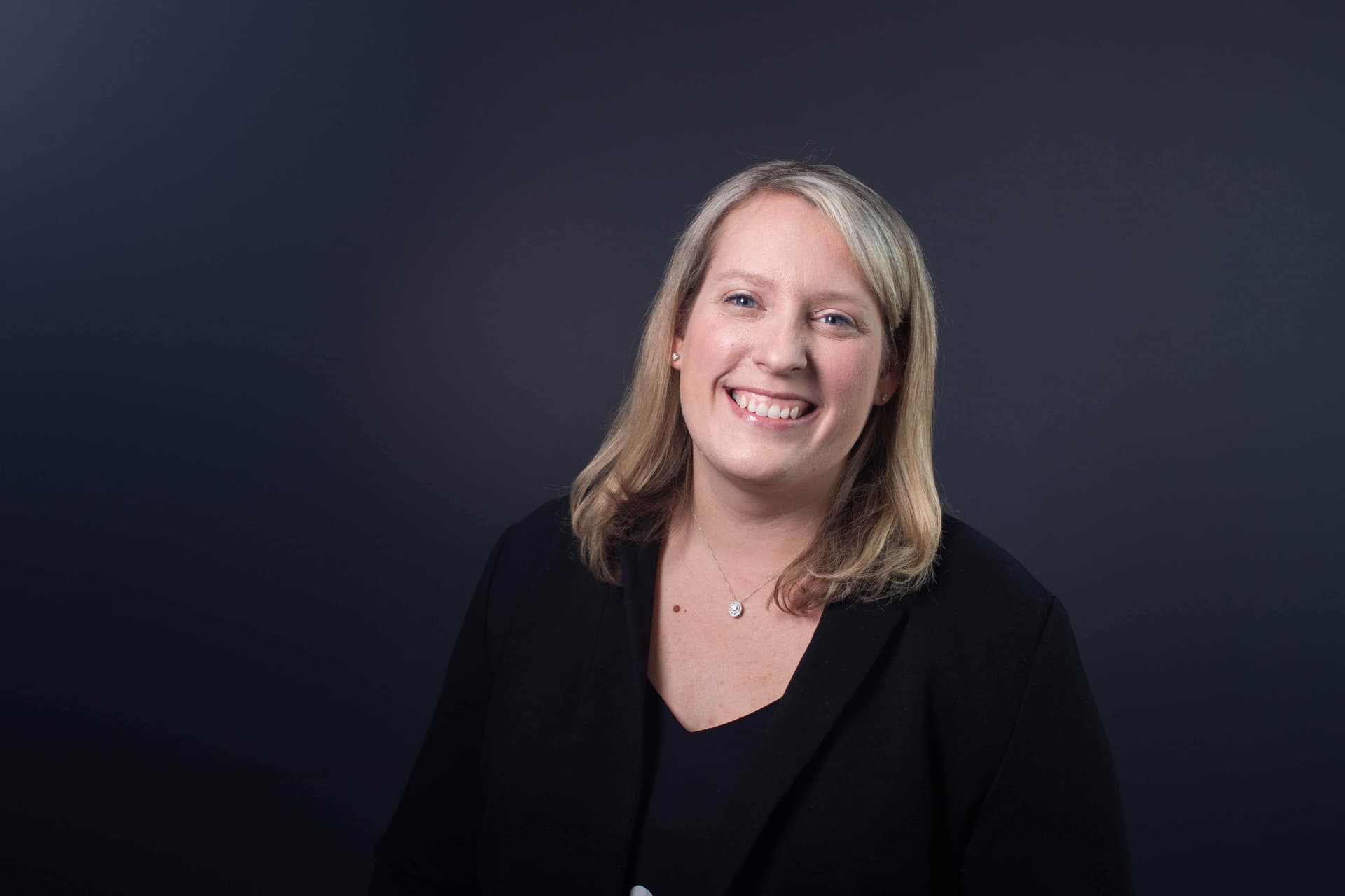 Professional woman smiling warmly in black blazer against dark background with soft studio lighting