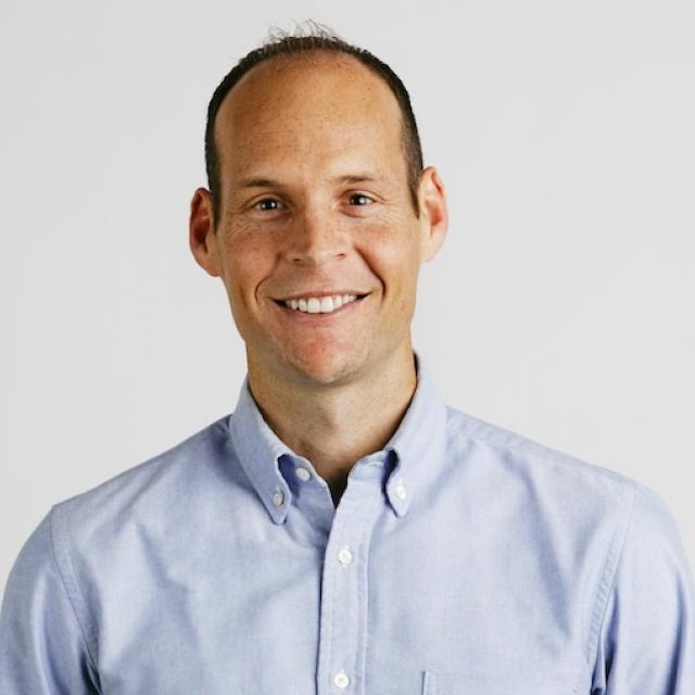 Professional headshot of a person wearing a light blue button-down shirt against a white background, smiling warmly at the camera