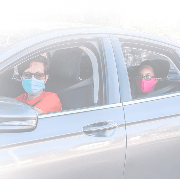 Two people wearing protective face masks sitting in a silver car, driver in blue mask and passenger in pink mask