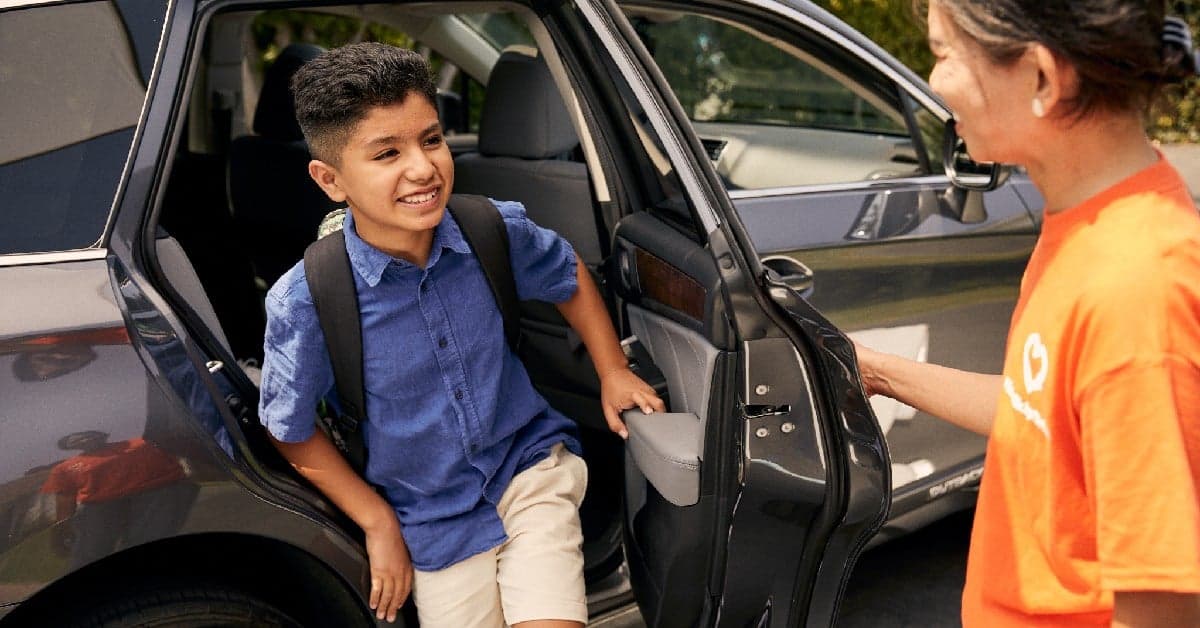 Young student with backpack getting into car, greeted by driver in orange company shirt
