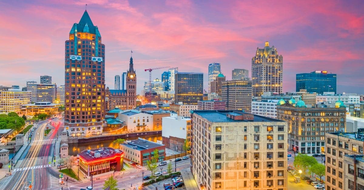 Downtown Milwaukee skyline with illuminated buildings against a pink and purple sunset sky, featuring historic and modern architecture