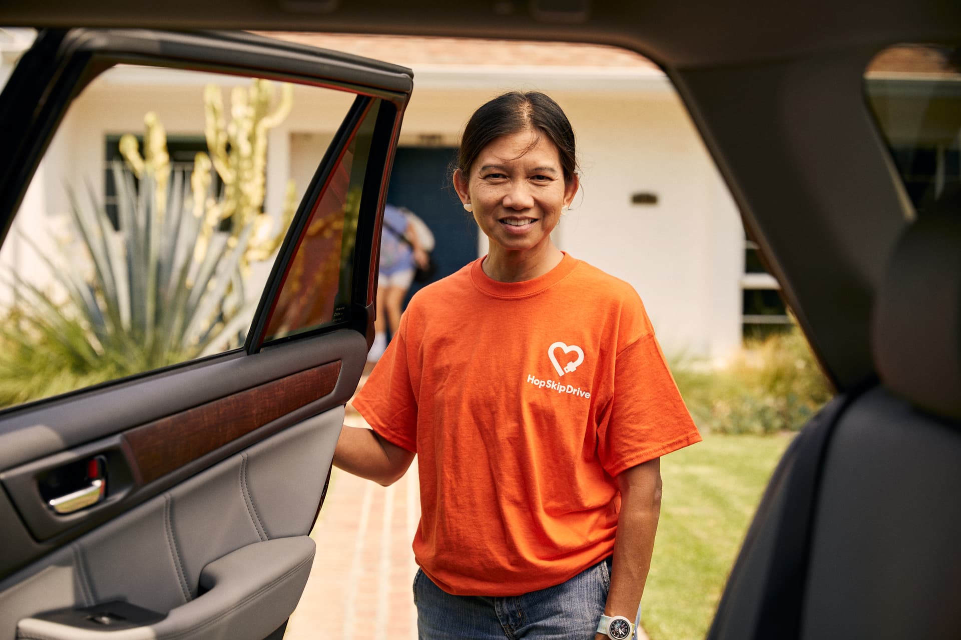Service provider in orange HopSkipDrive t-shirt standing by car door, smiling warmly at camera
