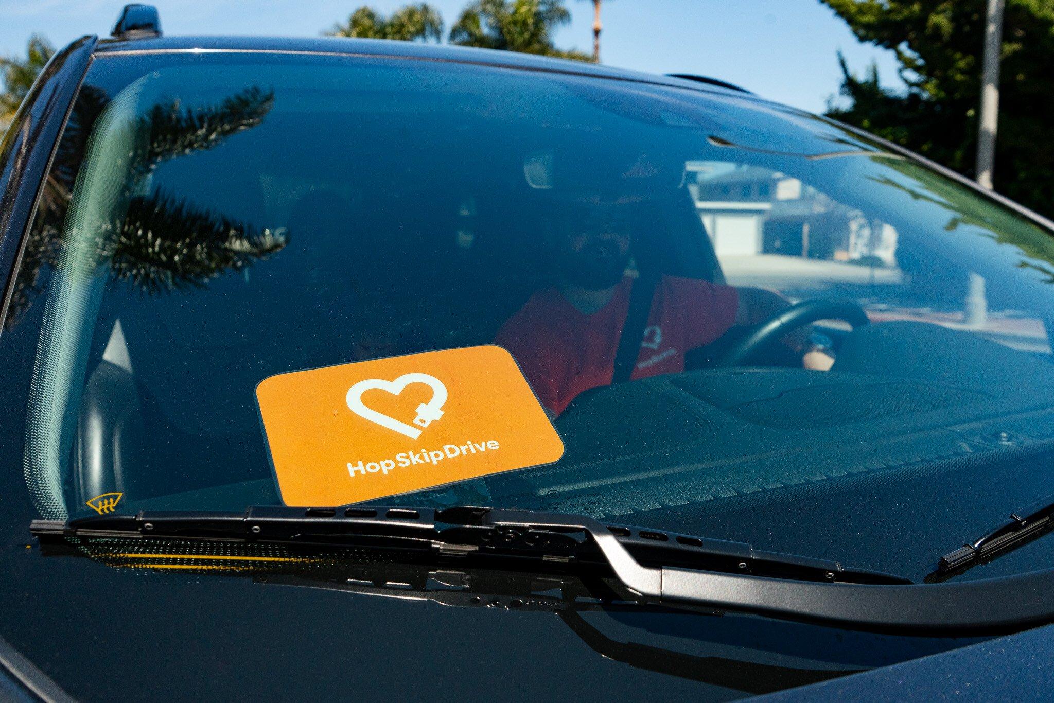Orange HopSkipDrive logo sticker with heart design displayed on a car windshield next to windshield wipers