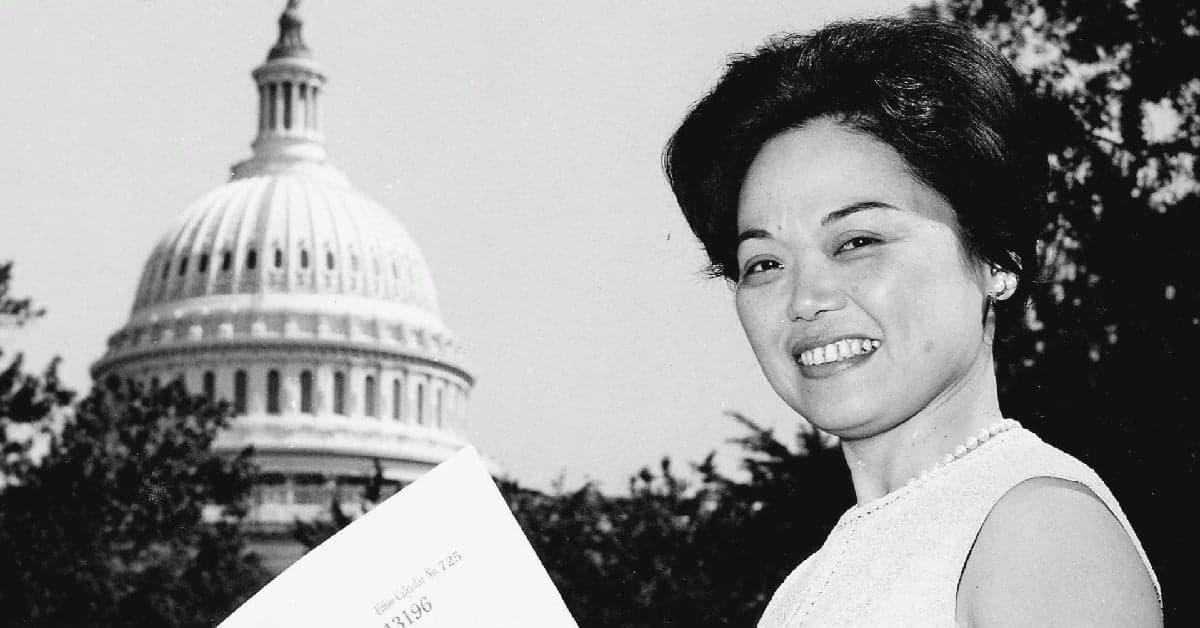 Woman in sleeveless white dress smiling while holding document, with US Capitol dome in background, black and white photograph