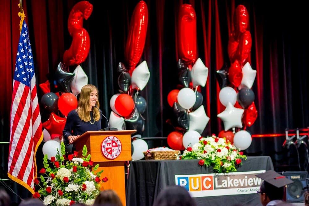 Graduation stage with speaker at podium, decorated with American flag, red and white balloons, and floral arrangements