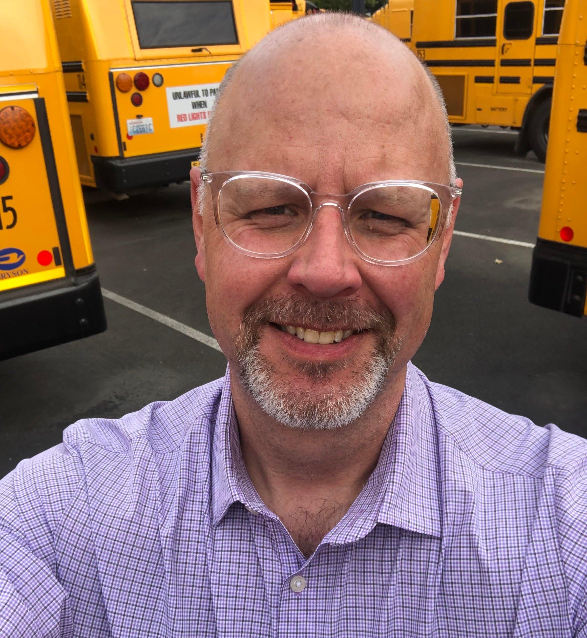 A smiling professional wearing glasses and a purple checkered shirt, with yellow school buses visible in the background