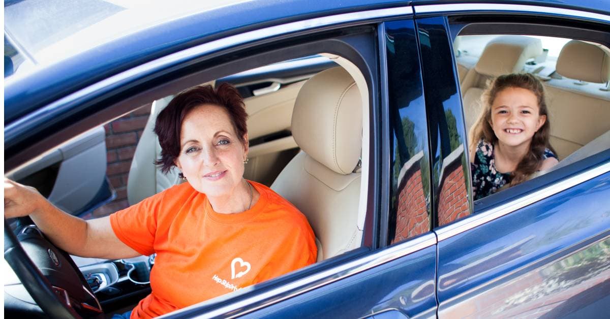 Adult volunteer in orange shirt driving car with young passenger smiling in back seat, both looking at camera