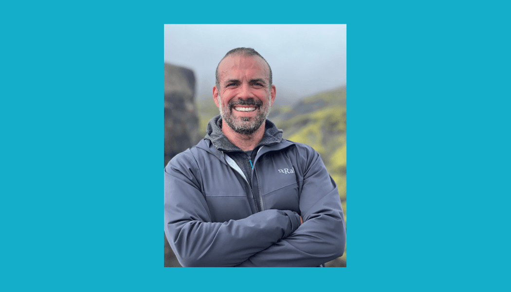 Portrait of a bearded man smiling warmly while wearing a grey Rab jacket outdoors with mountains in the background