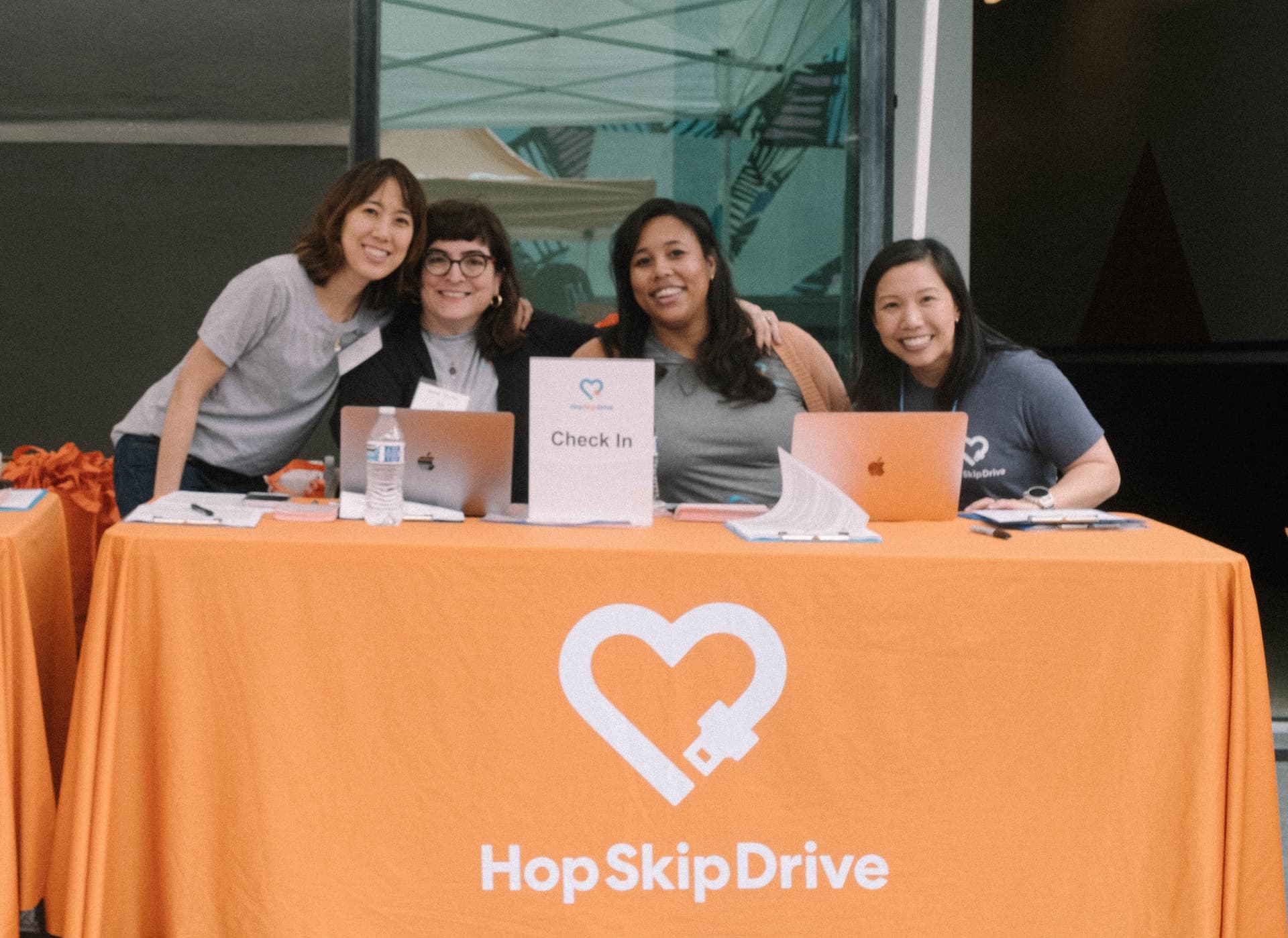 Four volunteers staffing a HopSkipDrive event check-in table with orange tablecloth, laptops, and registration materials