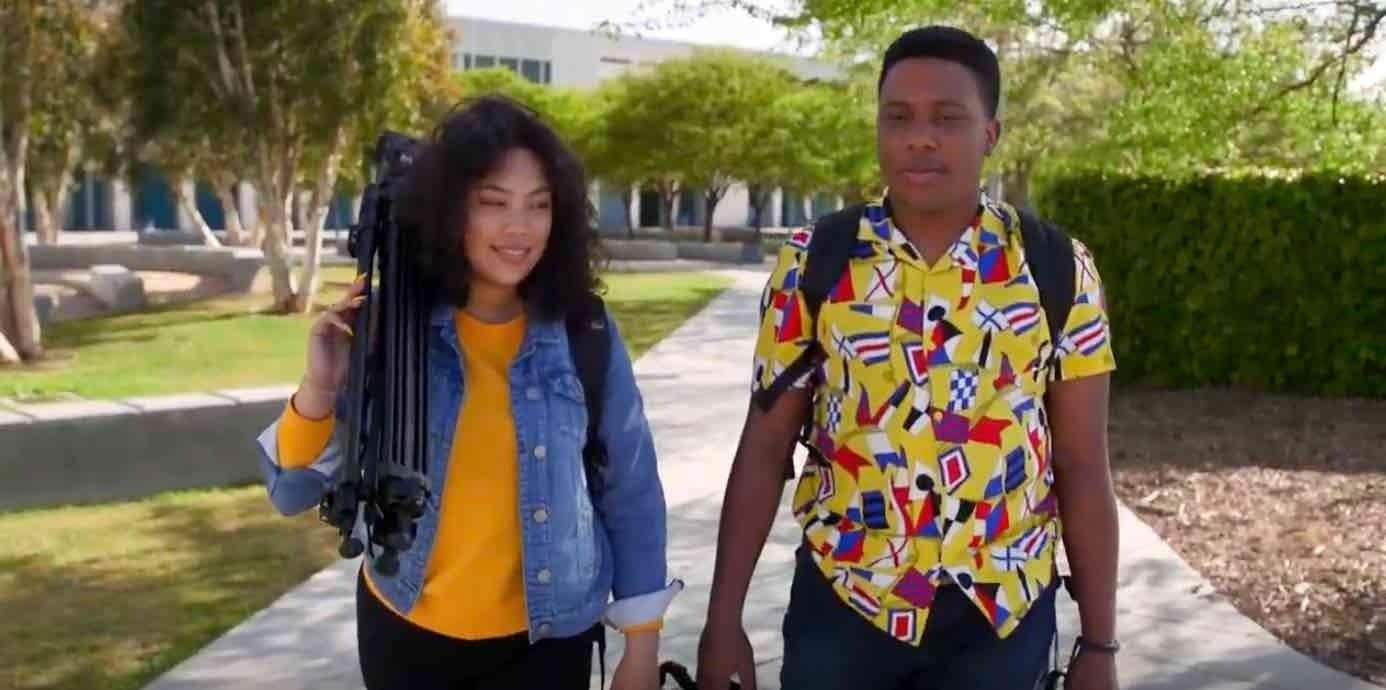 Two students walking on a tree-lined campus pathway, one carrying a tripod, wearing casual attire on a sunny day
