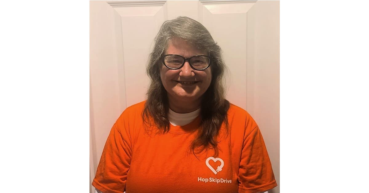 A person wearing glasses and an orange HopSkipDrive t-shirt with heart logo smiles at the camera against a white background