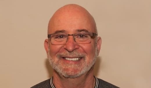 Professional headshot of a bald man with glasses and grey beard wearing a striped sweater, smiling warmly at camera