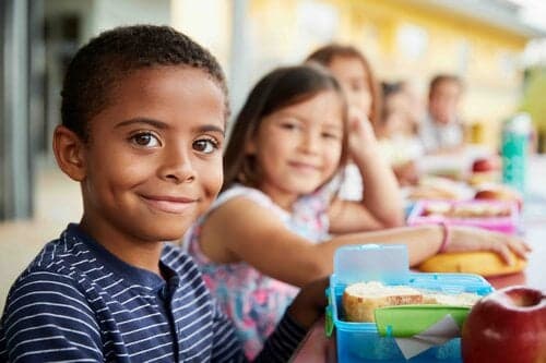 Elementary school students sitting at lunch table with packed lunches, smiling at camera