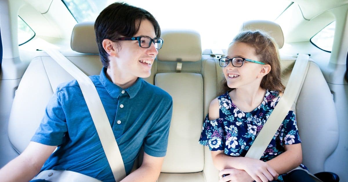 Two young people wearing glasses smiling at each other while sitting in car backseat, wearing blue polo and floral dress