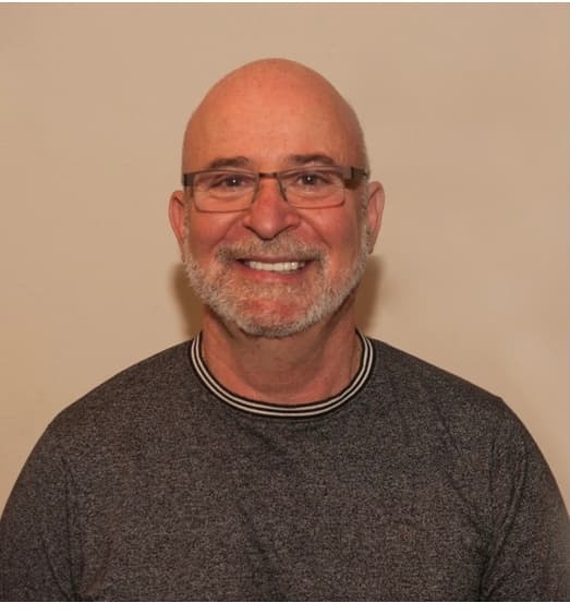 Friendly middle-aged man with glasses and gray beard wearing a gray sweater, smiling warmly at camera against neutral background