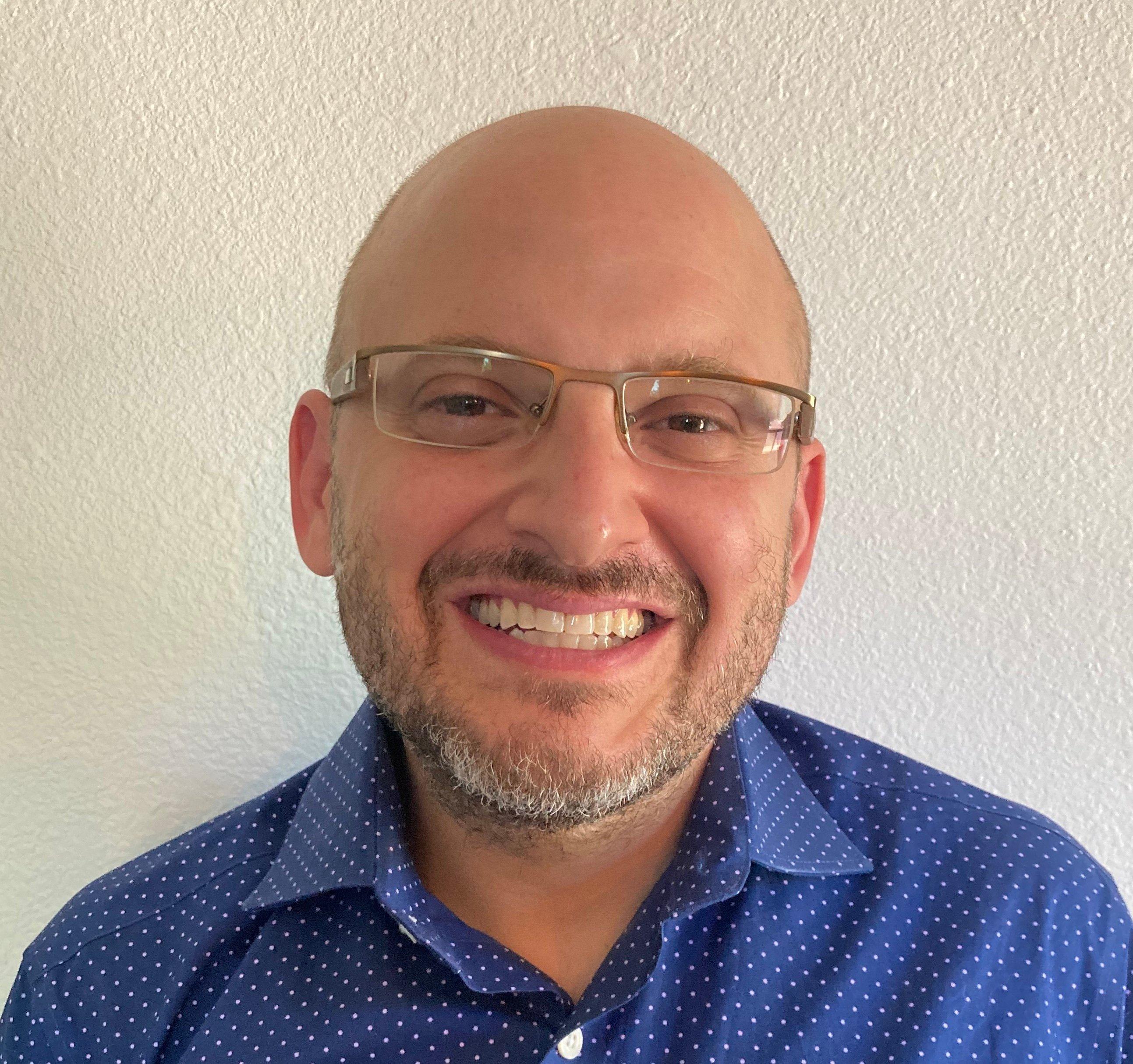 Professional headshot of person wearing glasses and blue polka dot shirt, smiling warmly at camera against light background