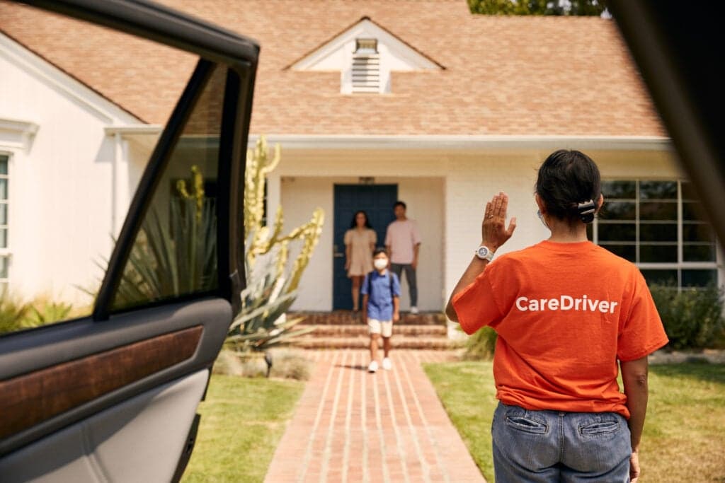 CareDriver in orange shirt waving goodbye to family standing at their house entrance after safe drop-off