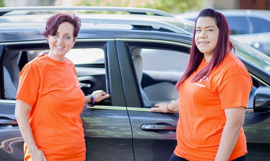 Two staff members wearing matching bright orange t-shirts standing by a dark-colored car in a parking lot