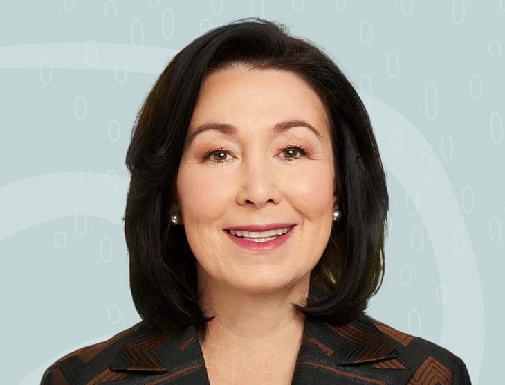 Professional headshot of an Asian woman in business attire smiling warmly, wearing pearl earrings against a light blue patterned background