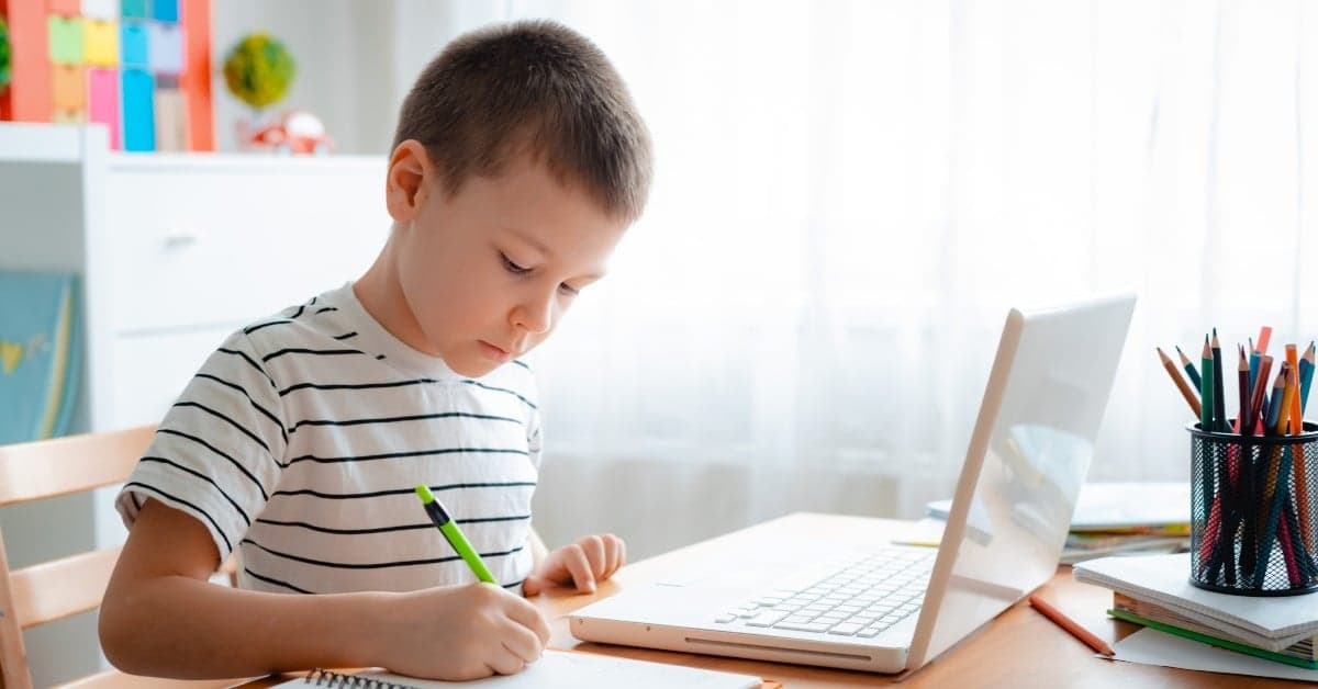 Young student in striped shirt writing while sitting at desk with laptop and school supplies in bright home study space