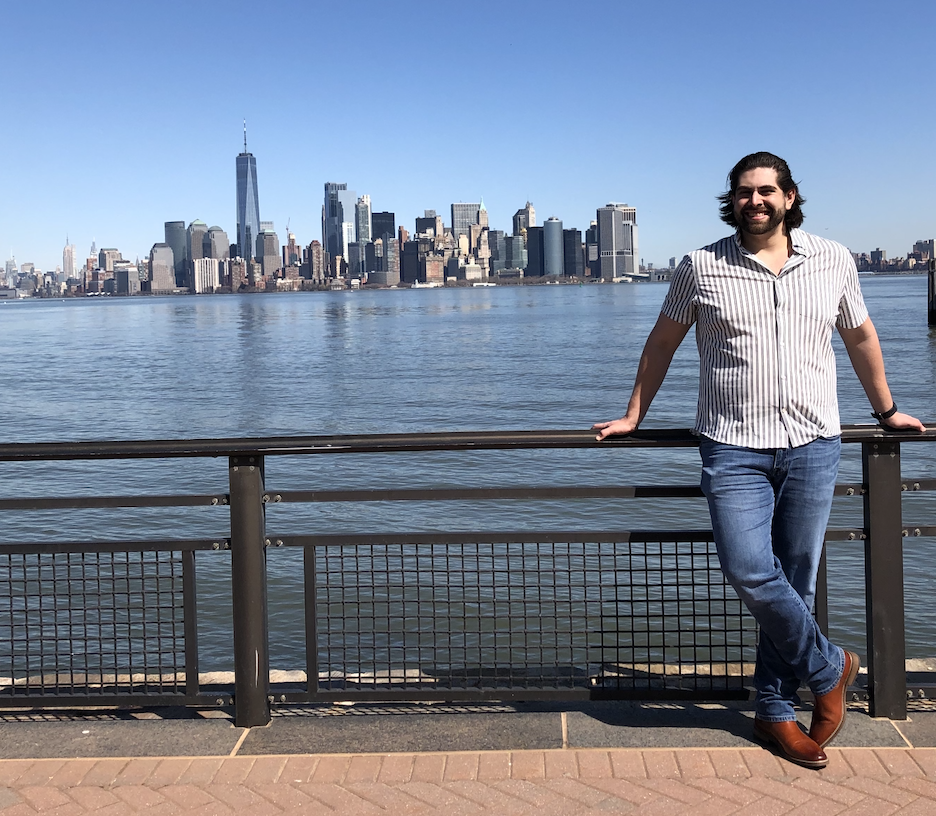 Tourist posing against Manhattan skyline view from waterfront, featuring One World Trade Center and downtown skyscrapers