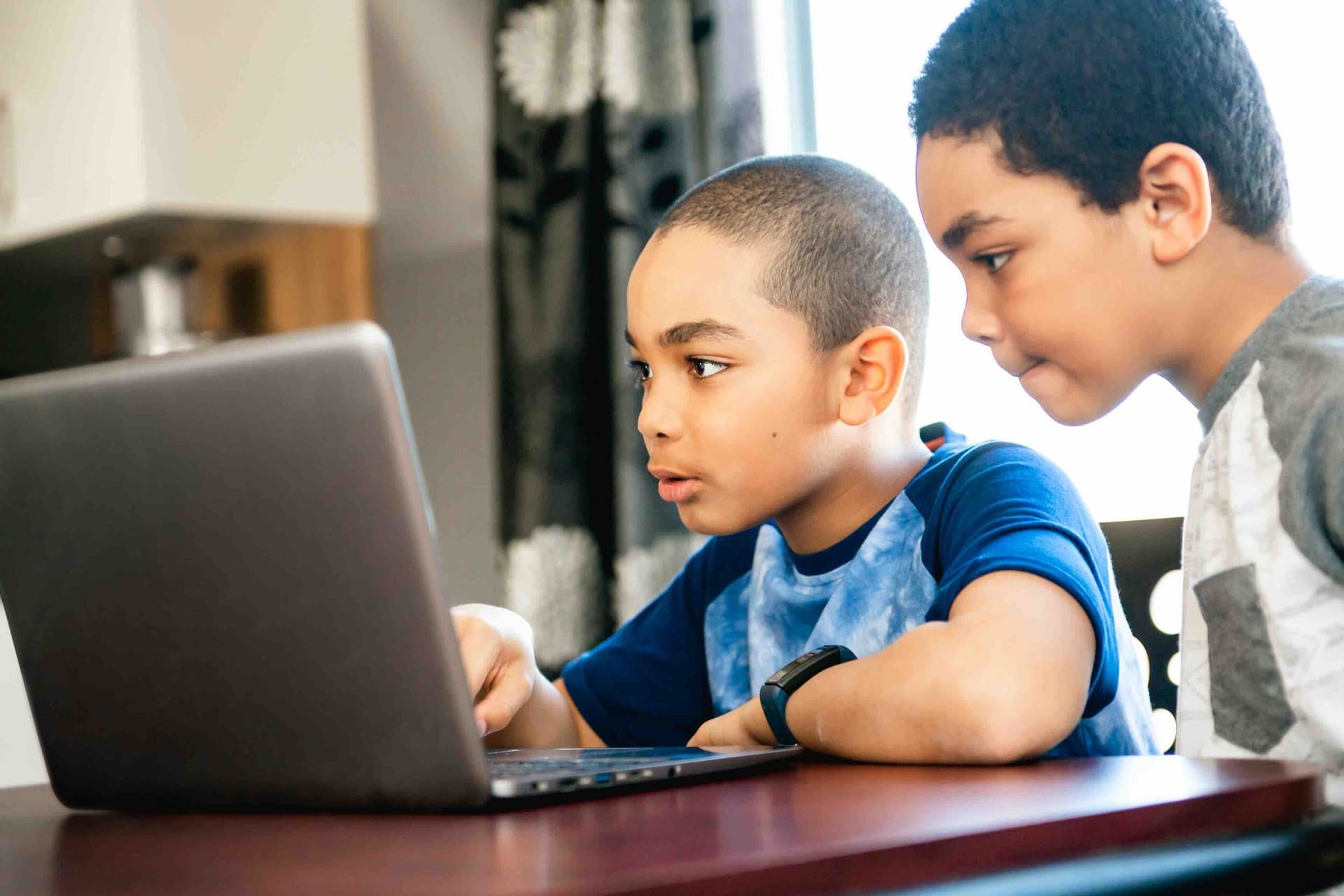Two young students working together at a laptop, focused intently on the screen in a home setting
