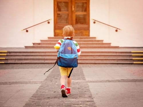 Young child wearing a blue backpack walking up to school entrance steps wearing yellow shorts and red shoes