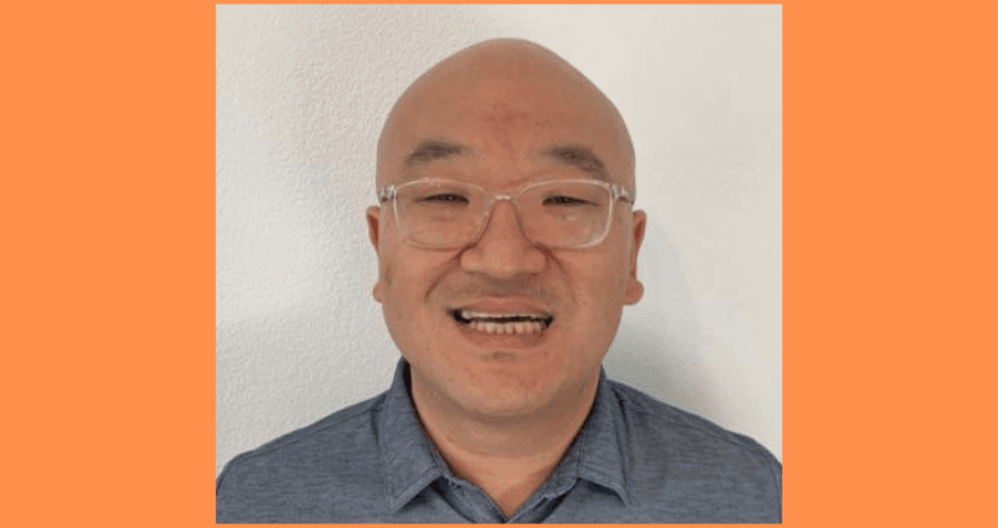 Professional headshot of a person wearing clear-framed glasses and a grey collared shirt, smiling warmly against a white wall