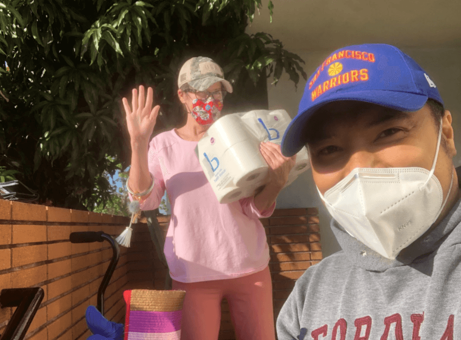 Two people wearing masks outdoors, one holding paper supplies and waving, another in Warriors cap taking a selfie