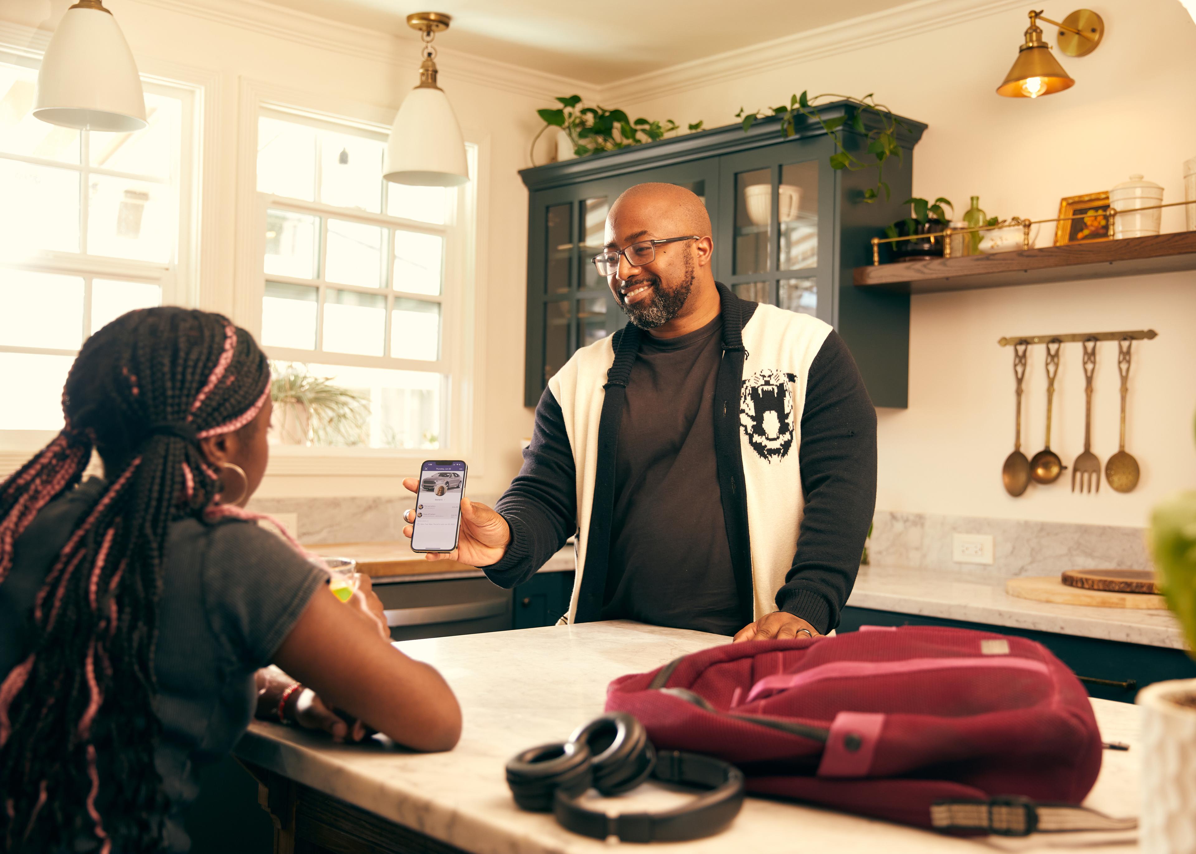Adult showing phone to young person across kitchen counter, with backpack and headphones on marble countertop in warm home setting