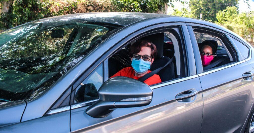 Two people wearing protective face masks sitting in a silver car, driver in red shirt and passenger in background wearing pink mask