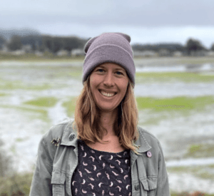Person wearing beanie and jacket smiling warmly against backdrop of natural wetland or tidal landscape
